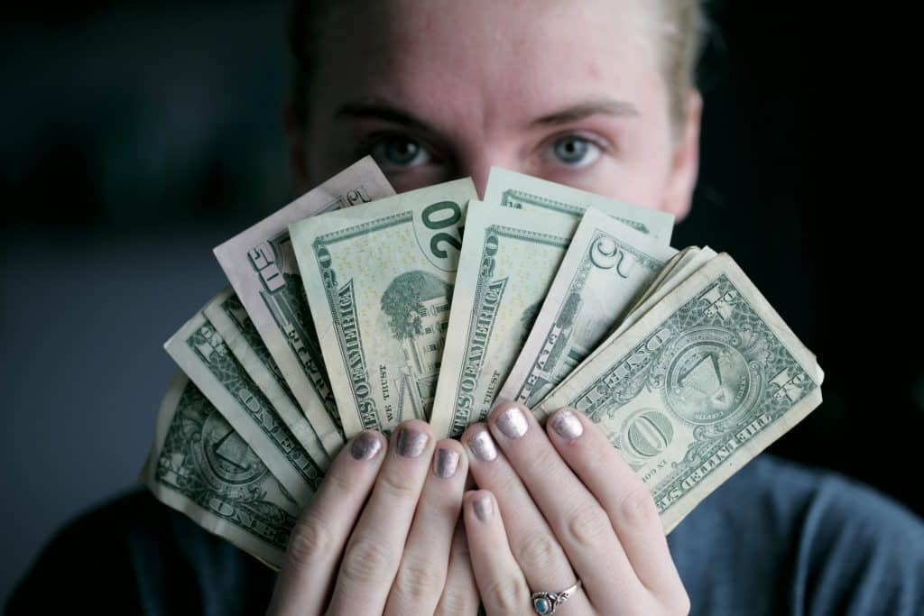 a white woman looks directly at the camera, holding a fan of green dollar bills that cover her face, leaving only her eyes visible.